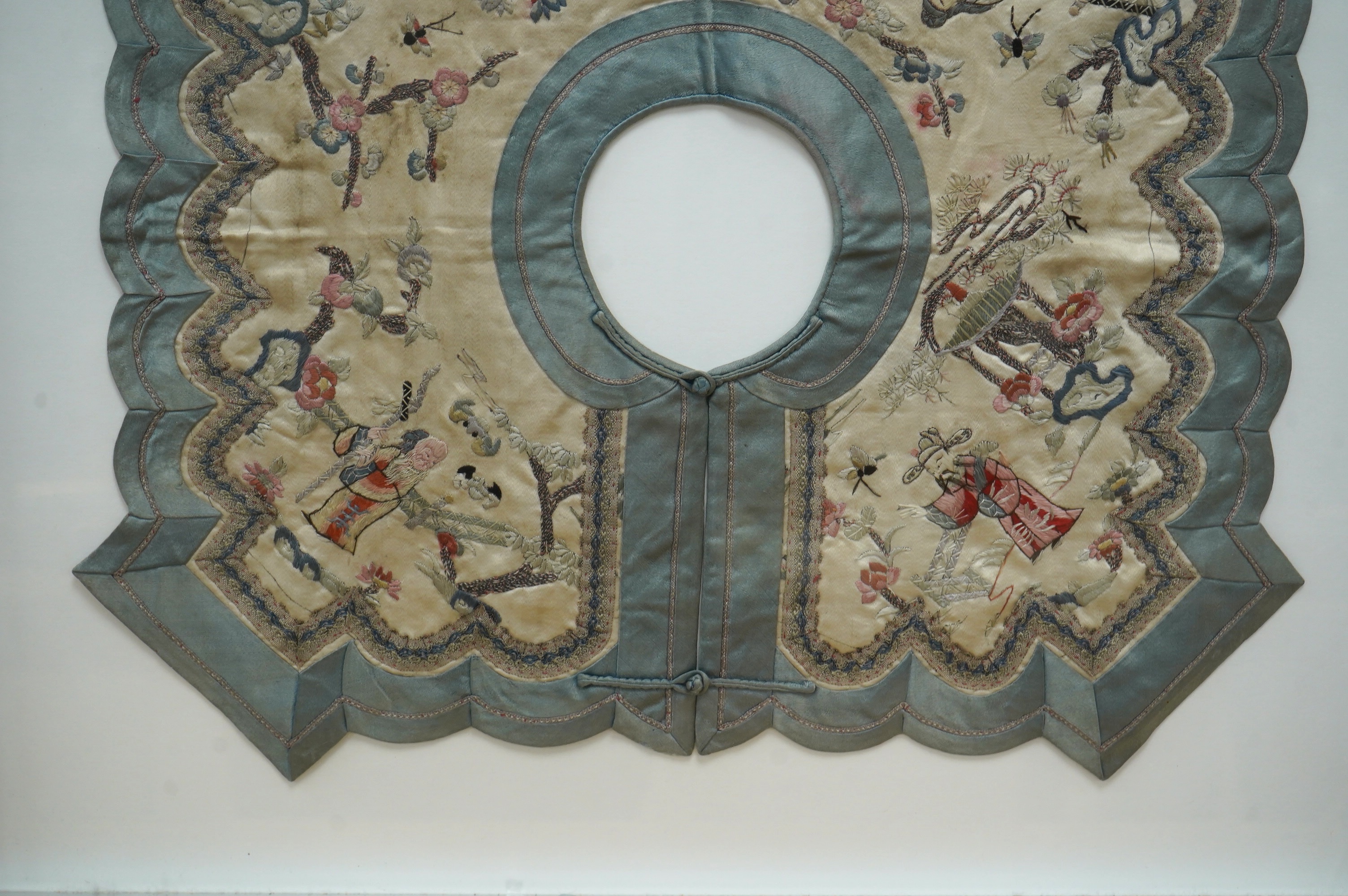 A framed Chinese ornate embroidered collar with scalloped edge, embroidered on cream silk, with polychrome silks, of figures, blossom, butterflies and auspicious symbols, the edges scalloped and braided with an outer blu